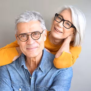 A smiling elderly couple posing for a photograph, with the woman resting her hand on the man's shoulder