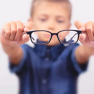 A boy with glasses is holding them in both hands and looking at them