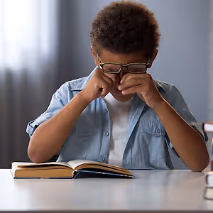 A young boy wearing glasses is sitting at a desk with his head in his hands, probably crying, while reading a book.
