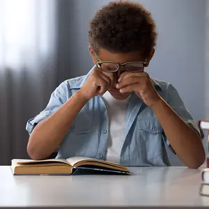 A young boy wearing glasses is sitting at a desk with his head in his hands, probably crying, while reading a book.