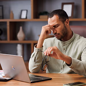A man working at a desk with a laptop, he is holding his head in his hand with glasses on his lap, he is wearing a green shirt.