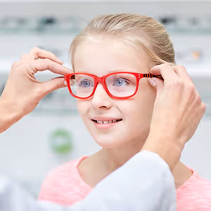 A young girl tries on red glasses while a doctor adjusts them for her