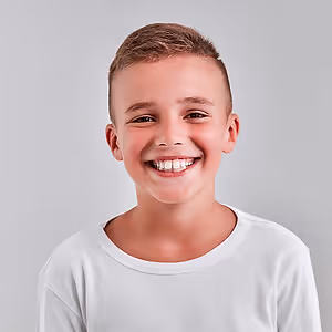 A young boy wearing a white t-shirt is smiling for a photograph against a gray background