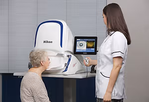 An elderly woman is sitting in front of a Nikon device in a medical room, a nurse is standing beside her, and the nurse is holding a remote control.