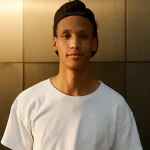 A man with a black cap and white t-shirt standing in front of a beige wall