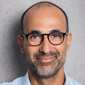 An adult man with a beard and glasses wearing a light blue shirt is smiling for a photo with a textured wall in the background