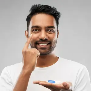 A man smiling and pointing to his eye with a blue contact lens in his hand