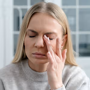 A blonde-haired woman with her eyes closed, gently touching her forehead with her fingers, possibly in a room with a white wall and glass window behind her.
