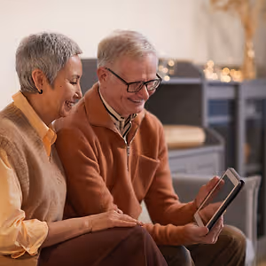 An elderly couple sits on a couch, the man wearing glasses and holding a tablet, and the woman wearing a beige sweater, both smiling and looking at the tablet