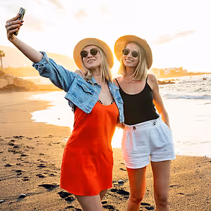 Two women are smiling on a beach and one is taking a selfie with her phone.