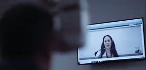 A woman wearing headphones is speaking into a camera on a monitor in a room with a blurry background
