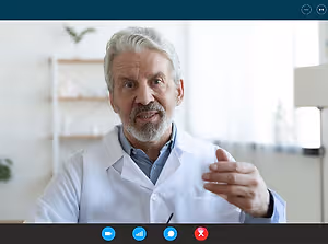 An elderly man wearing a white lab coat is speaking on a video call.