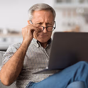 Elderly man sitting on a couch, holding his forehead with one hand, wearing glasses and looking at a laptop with a serious expression on his face.