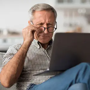 Elderly man sitting on a couch, holding his forehead with one hand, wearing glasses and looking at a laptop with a serious expression on his face.
