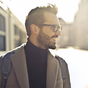 A man with a backpack wearing a coat and glasses standing in the city.