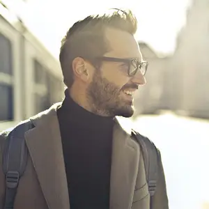 A man with a backpack wearing a coat and glasses standing in the city.