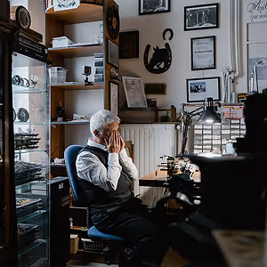An older man sits in a blue chair in front of a desk with an antique printing press, his hands covering his face as he gazes down. There is a wooden shelf and glass cabinet behind him, with a variety of items displayed, and framed pictures and a horseshoe on the wall.