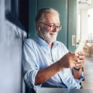 An older man with glasses is sitting on a chair and smiling while he is looking at his cellphone. He is wearing a bracelet and a necklace and has earphones on his ears. He is sitting in a restaurant with a table and a door behind him.