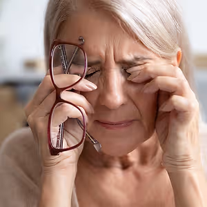an older woman holding her eyes and crying while holding a pair of glasses