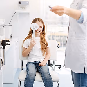 A young girl with long hair is holding a fan while sitting on a chair in a dental clinic with a dentist wearing a white lab coat standing in front of her.