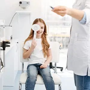 A young girl with long hair is holding a fan while sitting on a chair in a dental clinic with a dentist wearing a white lab coat standing in front of her.