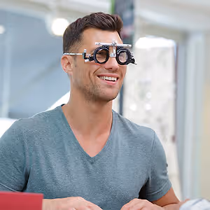 A smiling man wearing a gray V-neck shirt and a pair of glasses with a black frame and black lenses is looking at something while sitting in front of a glass wall.