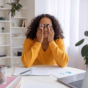 A woman in a yellow sweater is sitting at a desk, holding her glasses and covering her eyes.