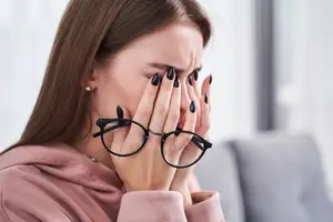 A woman wearing glasses appears to be in pain while covering her face with her hands.