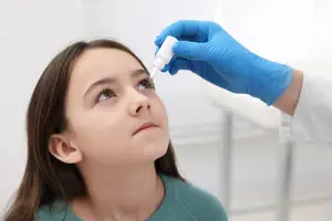 A doctor wearing blue gloves is administering eye drops to a young girl with brown hair and green sweater