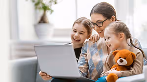 A woman with two young girls is sitting on a couch, one of the girls is smiling, and the other is holding a teddy bear while looking at a laptop