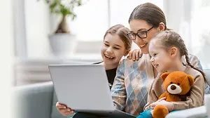 A woman with two young girls is sitting on a couch, one of the girls is smiling, and the other is holding a teddy bear while looking at a laptop