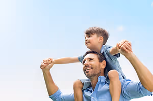 A smiling man wearing a blue shirt is holding a young boy on his shoulders under a clear blue sky.