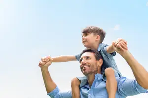 A smiling man wearing a blue shirt is holding a young boy on his shoulders under a clear blue sky.