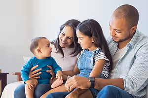 A smiling family of three, a man, a woman, and a young girl sitting on a couch with a baby boy on the woman's lap