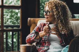 A woman wearing glasses is sitting on a chair holding a cup and smiling while looking to the left