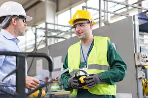 Two workers in a factory, one is talking to another while wearing safety gear.