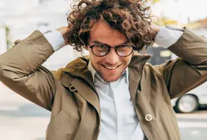 A man with curly hair wearing glasses and a coat is smiling and posing for a photo.