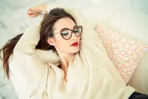 Woman lying on a couch with a white blanket and a patterned pillow behind her head wearing glasses and a beige sweater
