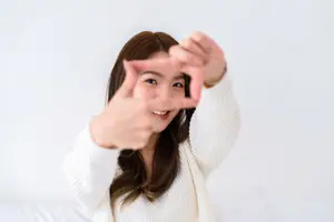 A woman wearing a white sweater is smiling and making a heart shape with her hands