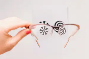 A person wearing glasses is holding a pair of glasses with spiral design on the lenses in front of a white wall.