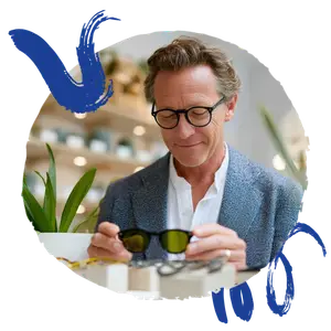A man wearing glasses is adjusting a pair of sunglasses with green lenses while smiling and looking at them
