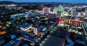 An aerial view of a vibrant city with numerous buildings and skyscrapers, many of which have neon lights, set against the backdrop of a mountain range at dusk.