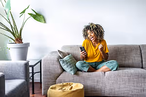 A woman is sitting on a couch and seems to be laughing while looking at her cell phone.