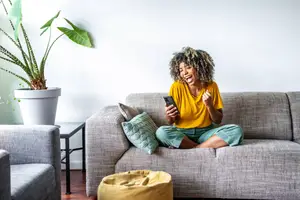 A woman is sitting on a couch and seems to be laughing while looking at her cell phone.