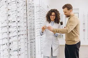 A man and a woman standing in front of a glass wall displaying many eyeglasses on display stands, the man is holding an eyeglass in his hand, and the woman is smiling