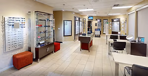 An optical store with a variety of eyewear on display, including glasses and sunglasses, arranged on shelves and tables, with a comfortable seating area, and digital signage displaying promotional offers. The space is well-lit with ceiling-mounted lamps and features a clean, modern design with beige walls and tiled flooring. The store also includes a monitor on the wall for displaying information or advertisements.