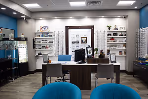 An office with wooden desks, blue chairs, and shelves displaying various items, including a monitor, potted plants, and picture frames. The room has a blue wall, a glass case with items, and a ceiling with lights.