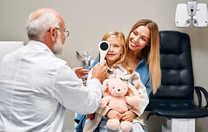 A doctor is examining a young girl's eye with a flashlight while a woman and a stuffed animal watch nearby.