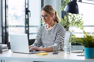 A woman with glasses working on a laptop at her desk.