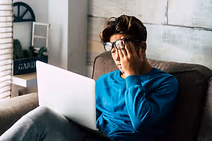 A young man wearing glasses and a blue long-sleeve shirt sits on a couch with his laptop, looking tired and holding his head.